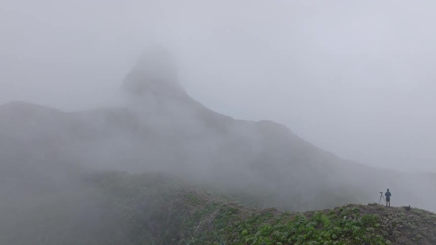 A slow aerial shot pushes in on a lone photographer with a tripod standing on a misty, green mountain ridge