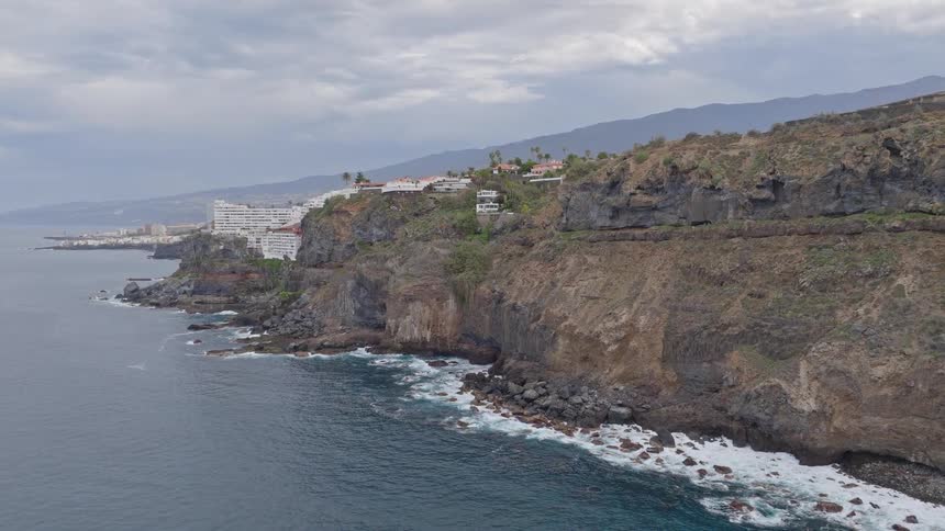 An aerial view shows a coastal town perched on rugged cliffs as ocean waves crash against the shore on a cloudy day