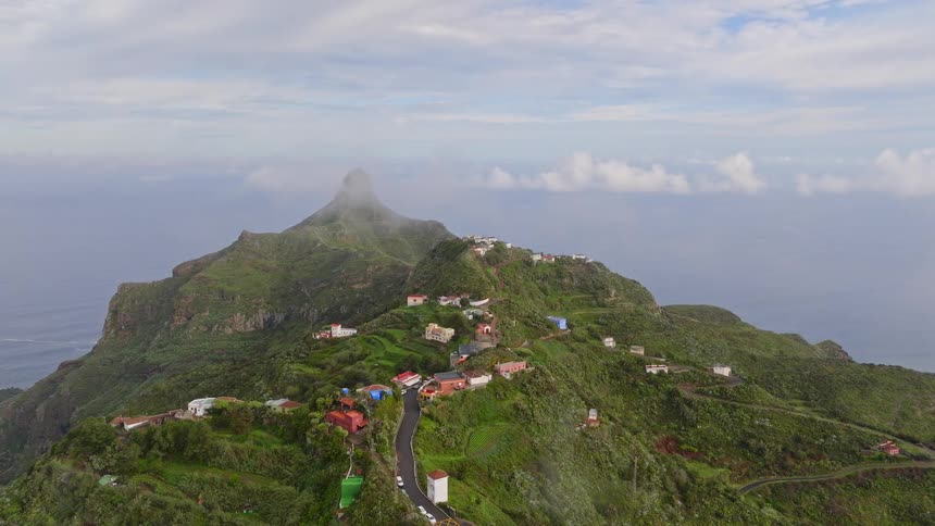 Thick fog and clouds dissipate to reveal a scenic village nestled high on a lush green mountain overlooking the ocean