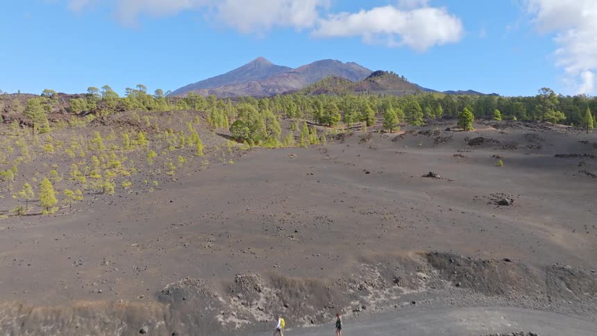 An aerial shot follows two hikers walking along a desolate trail, revealing the vast volcanic landscape of Teide National Park in Tenerife