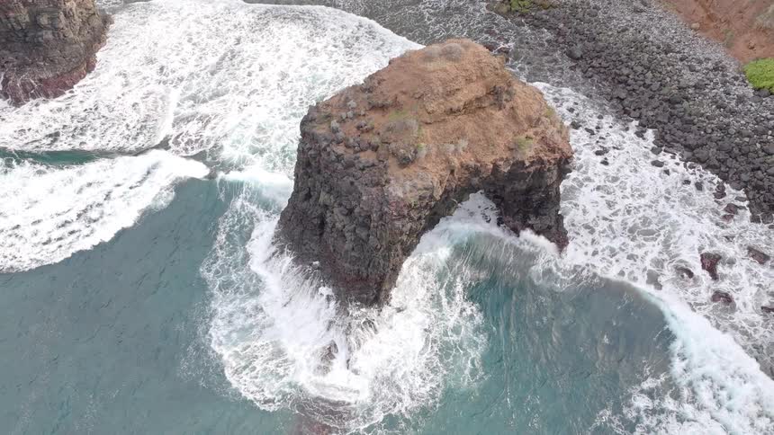 Slow motion aerial view ascending over a volcanic rock arch with waves crashing on the rugged coast