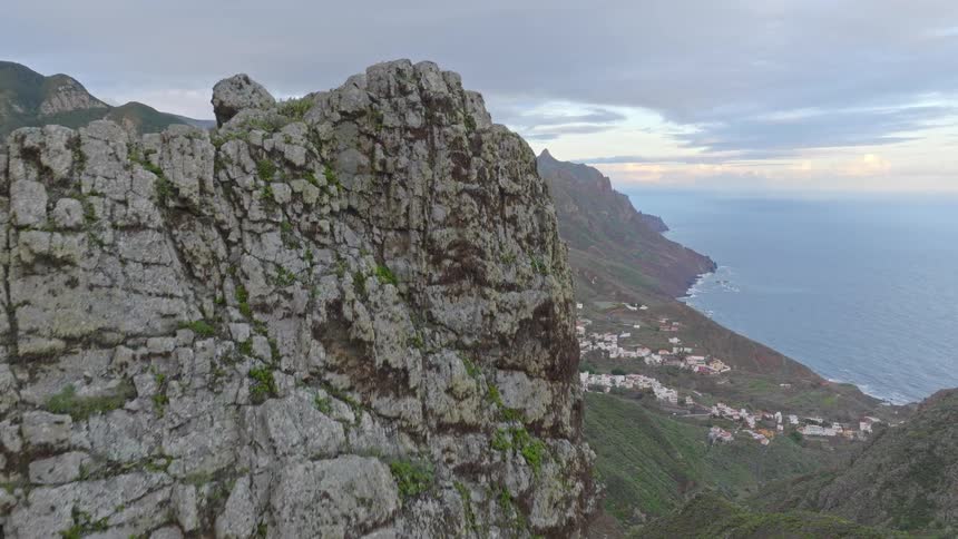 A slow aerial ascent over a rocky peak unveils a breathtaking view of a lush mountain range and a coastal village under a cloudy sky