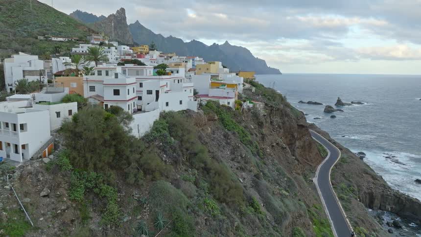 Aerial footage reveals a charming village perched on a dramatic cliffside overlooking the vast ocean and a winding coastal road
