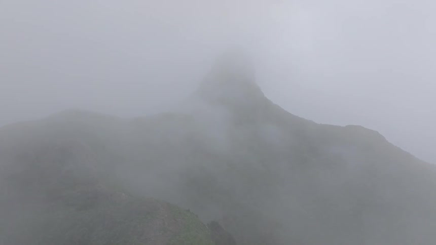 An aerial drone shot slowly pulls back over a green mountain peak completely enveloped in a thick, mysterious fog
