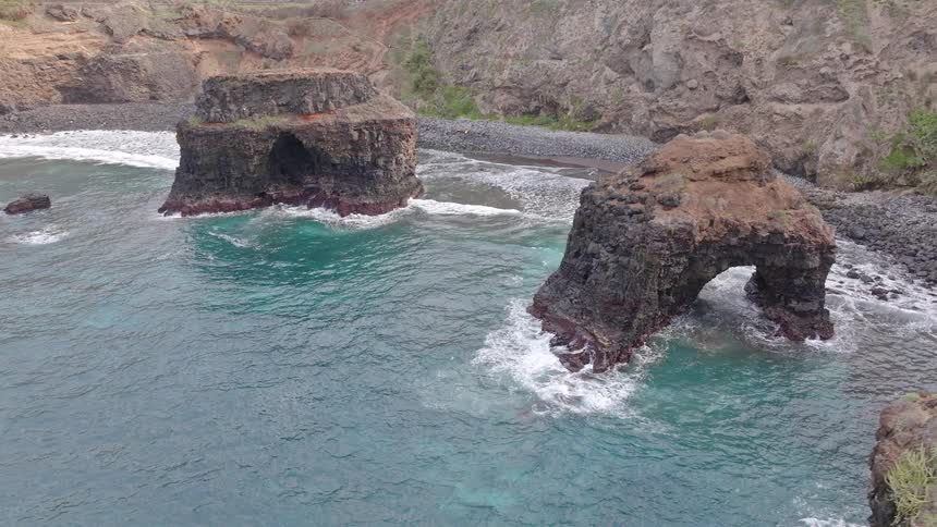 An aerial view pulls back to reveal stunning volcanic sea stacks and a natural rock arch in a secluded turquoise ocean cove