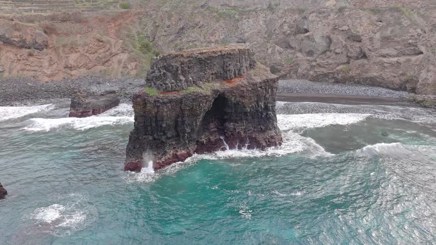 An aerial view slowly pulls back from a dramatic sea stack with a natural arch as turquoise ocean waves crash on a rugged coastline