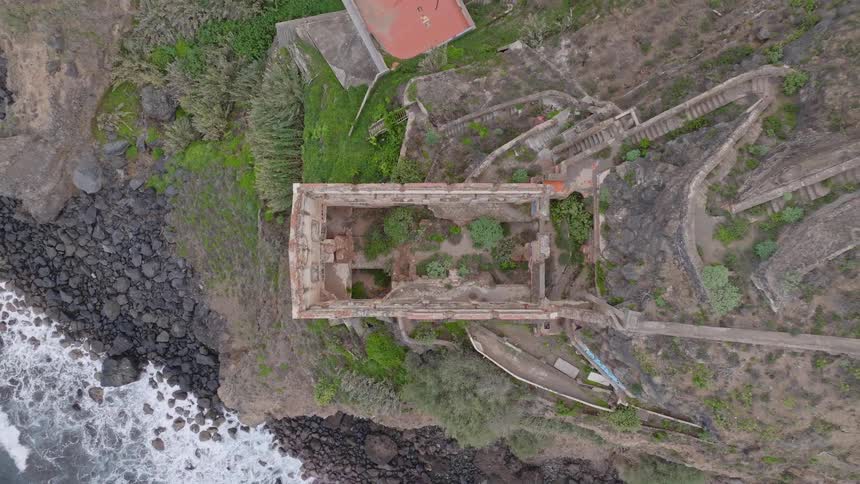 Aerial top-down view slowly ascends from a ruined, roofless building, revealing its location on a rocky cliff beside the ocean