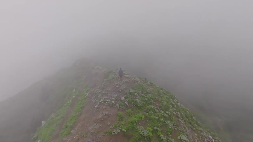 Aerial view of a lone photographer standing on a lush, foggy mountain peak next to a camera on a tripod