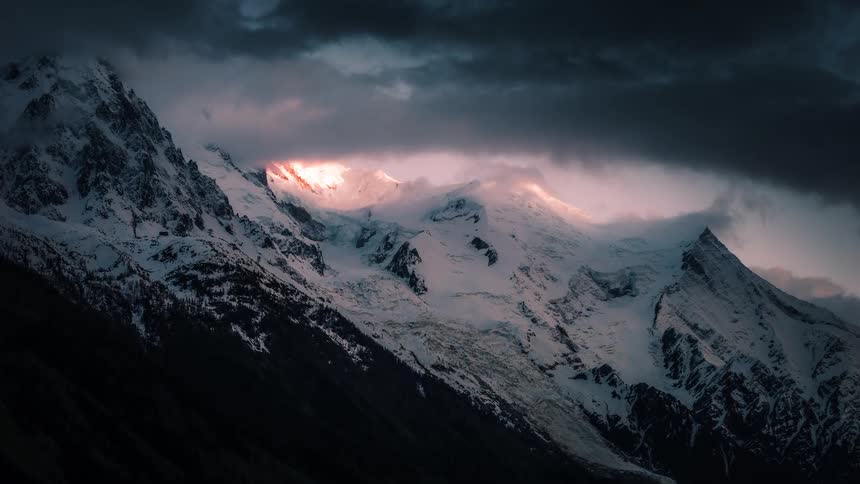 A dramatic time-lapse captures the setting sun's last light on a snowy mountain range under stormy skies