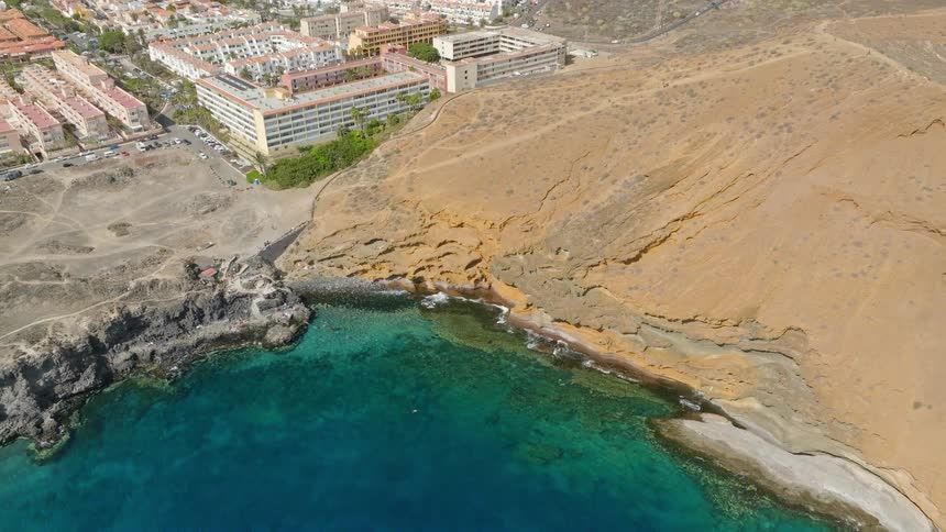 A stunning top-down aerial shot reveals the vibrant turquoise ocean meeting a unique golden-brown coastline, before pulling back to show the adjacent resort town