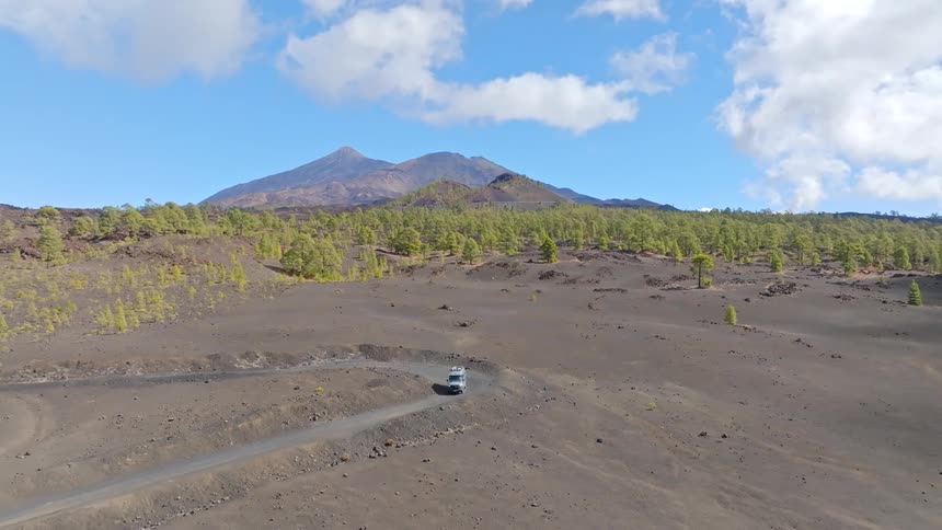 An epic aerial shot follows a camper van driving on a remote road through a vast volcanic landscape with mountains and a pine forest