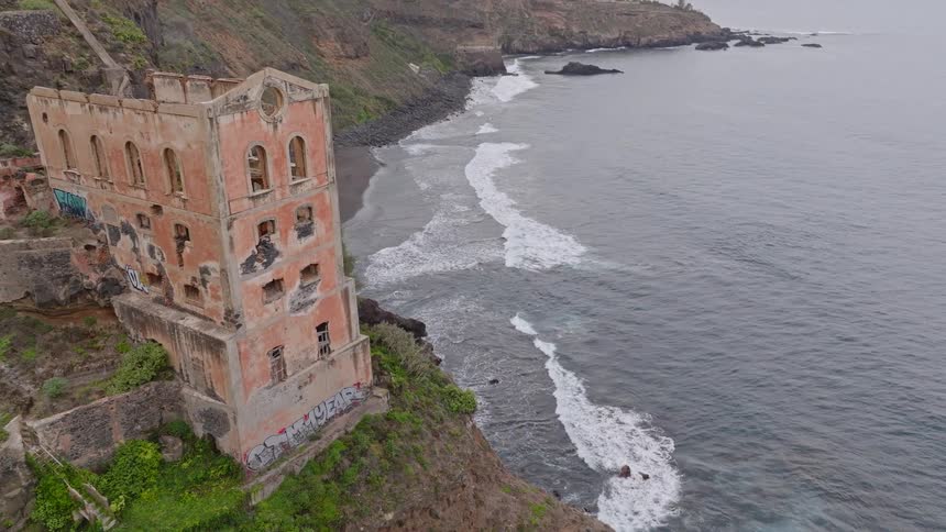 Eerie aerial shot descends to reveal the crumbling ruins of an abandoned building on a rugged cliffside overlooking the ocean