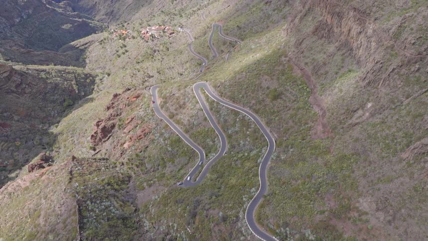 An aerial view shows cars navigating a winding serpentine road through a rugged mountain landscape, revealing a vast canyon and the ocean in the distance