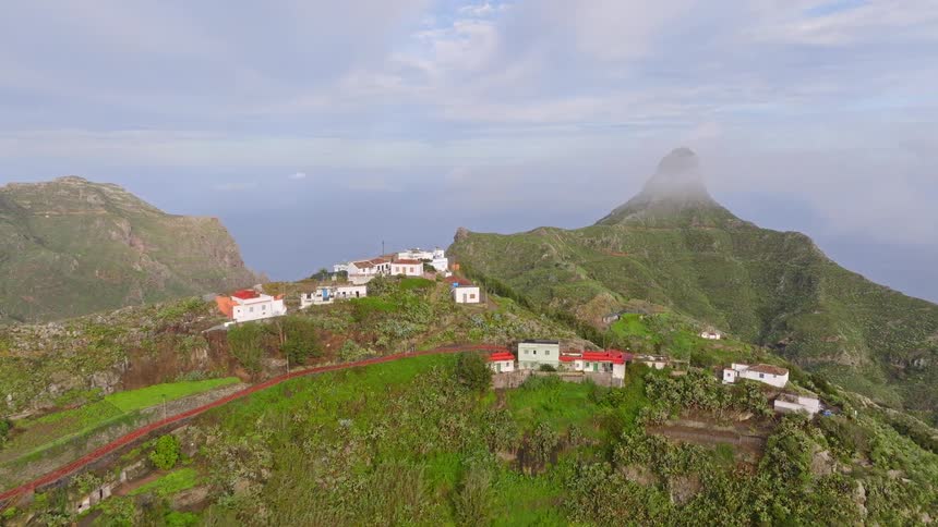 A slow aerial ascent over a charming village on a verdant mountain ridge with a dramatic, misty peak and ocean in the background