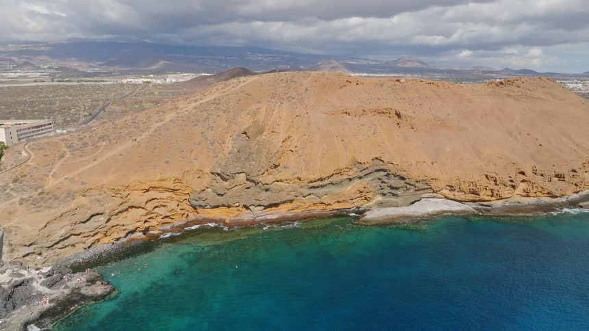 A stunning aerial pull-back shot reveals the volcanic landscape of Montaña Amarilla and the crystal-clear ocean in Tenerife