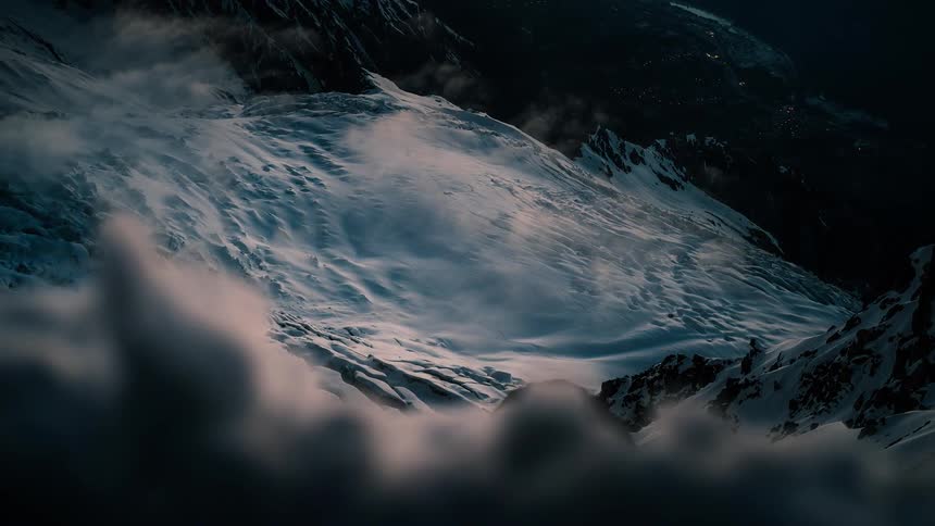 Cinematic timelapse of powerful winds sweeping snow across a dark mountain glacier as clouds roll through