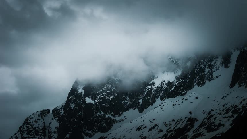 A dark and moody time-lapse shows storm clouds swirling over a dramatic, snow-covered mountain peak