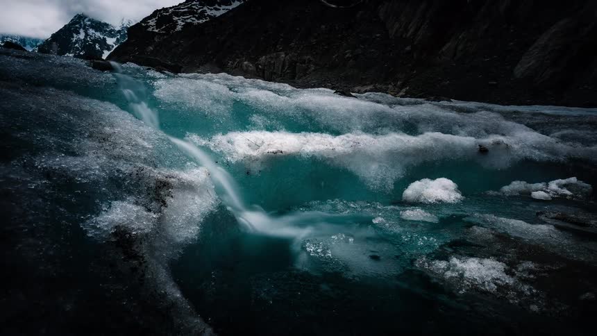 A dramatic time-lapse with a slow zoom out reveals a vibrant turquoise meltwater stream carving a path through a glacier in a stark mountain valley