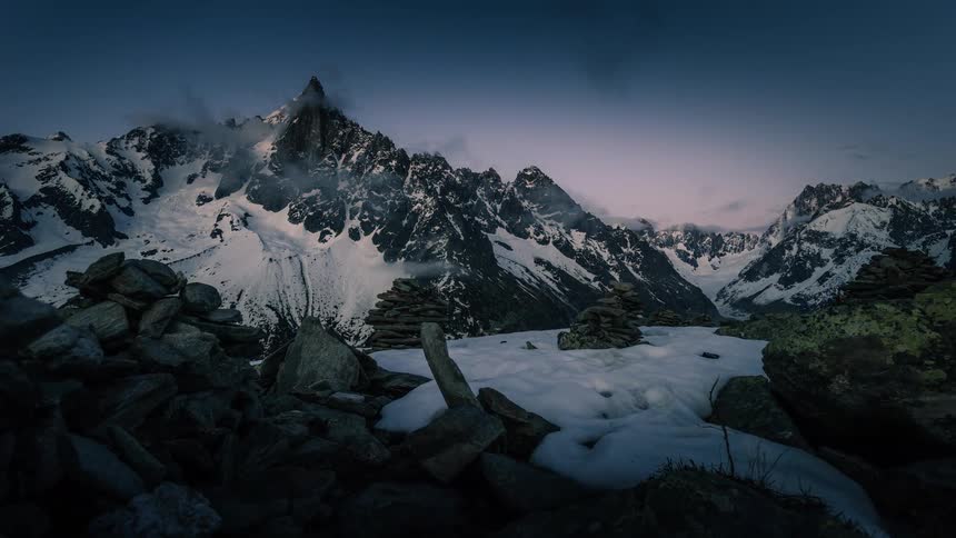 An epic day-to-night timelapse of clouds swirling around majestic, snow-covered mountain peaks with stone cairns in the foreground