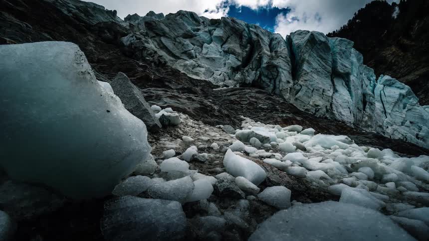 A dramatic low-angle timelapse reveals the immense scale of a rugged glacier wall with chunks of ice in the foreground