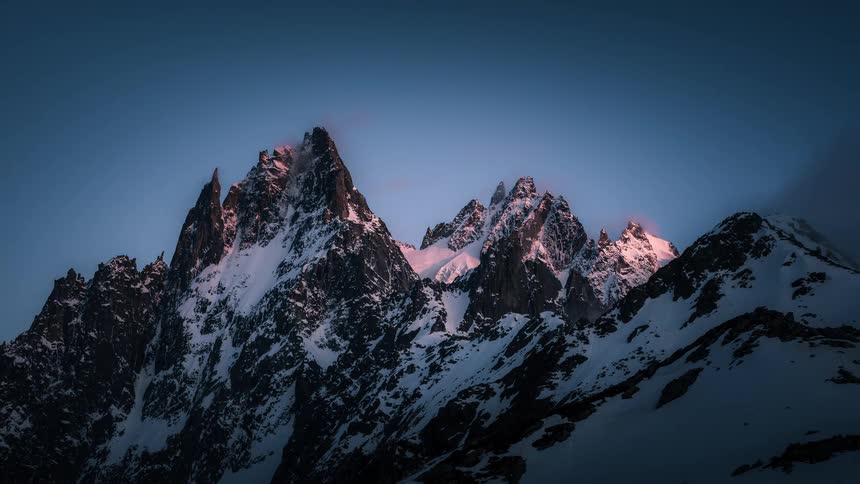 A beautiful time-lapse of clouds swirling around majestic, snow-covered mountain peaks as they are illuminated by the warm light of a setting sun