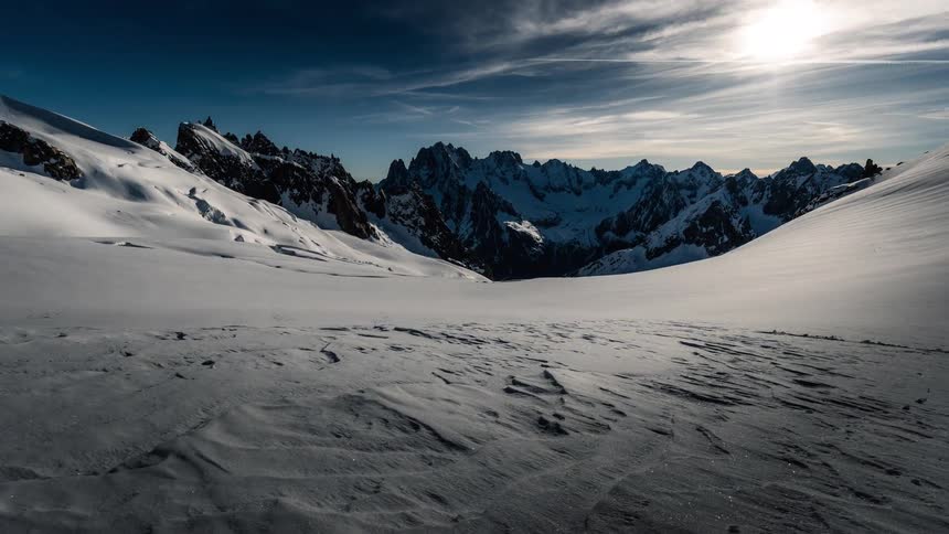 A dramatic timelapse shows shadows moving across a vast, frozen glacier valley surrounded by jagged, snow-covered mountain peaks