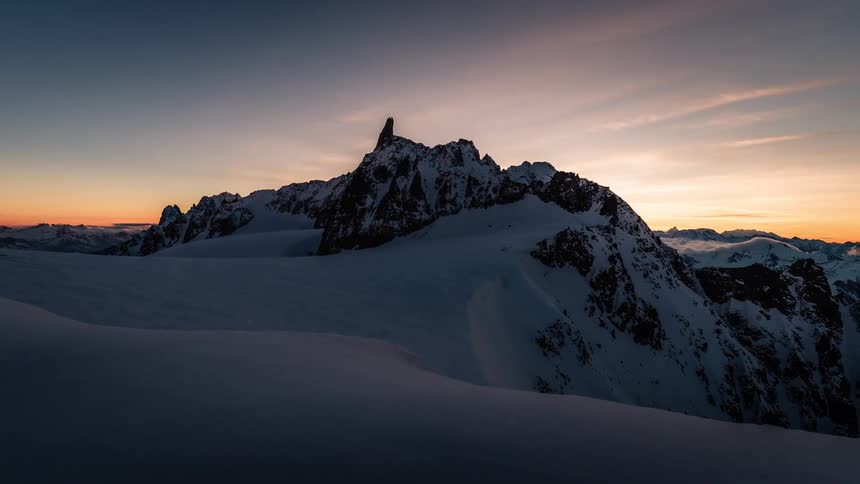 A breathtaking timelapse captures the sun rising over a vast and silent snow-covered alpine landscape, casting a warm glow on the jagged peaks