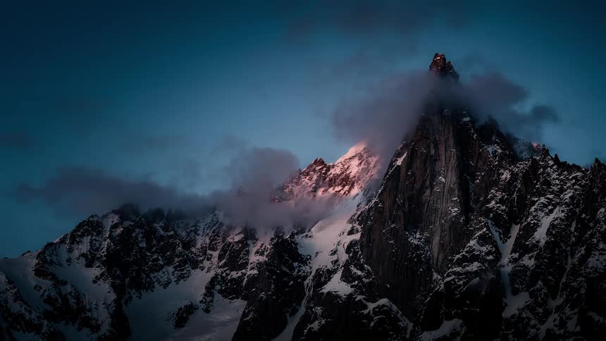 Time-lapse of clouds swirling around a majestic, snowy mountain peak as the warm sunset glow fades to dusk