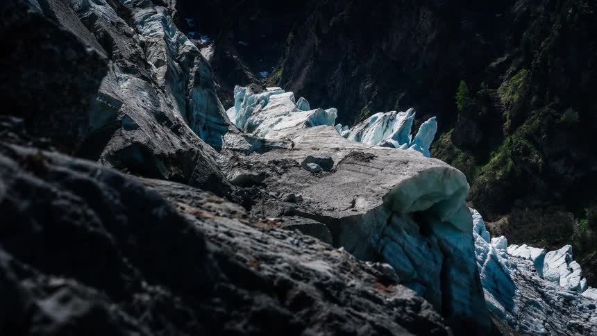 A dramatic dolly-out timelapse reveals the immense scale of a glacier in a mountain pass, with sunlight and shadows playing across the icy surface