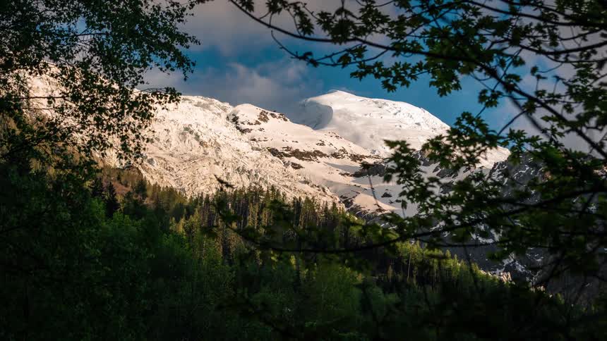 Sunlight and shadows dance across a snow-covered mountain peak in a beautiful time-lapse framed by verdant forest leaves