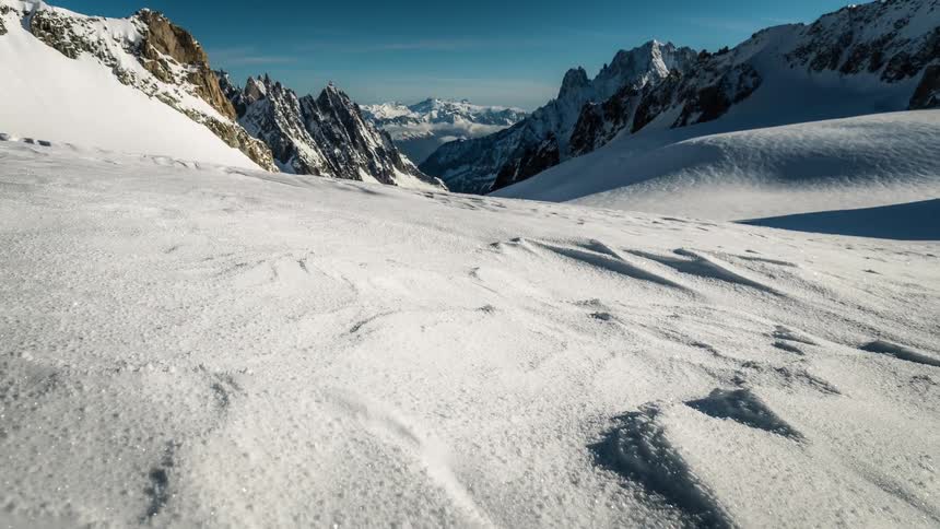 Slowly pulling back over pristine snow to reveal a vast, breathtaking alpine valley surrounded by jagged peaks under a clear blue sky