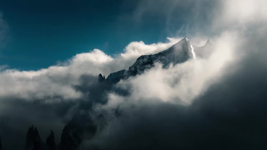 A dramatic time-lapse of thick clouds swirling and revealing a snow-covered mountain peak under a deep blue sky