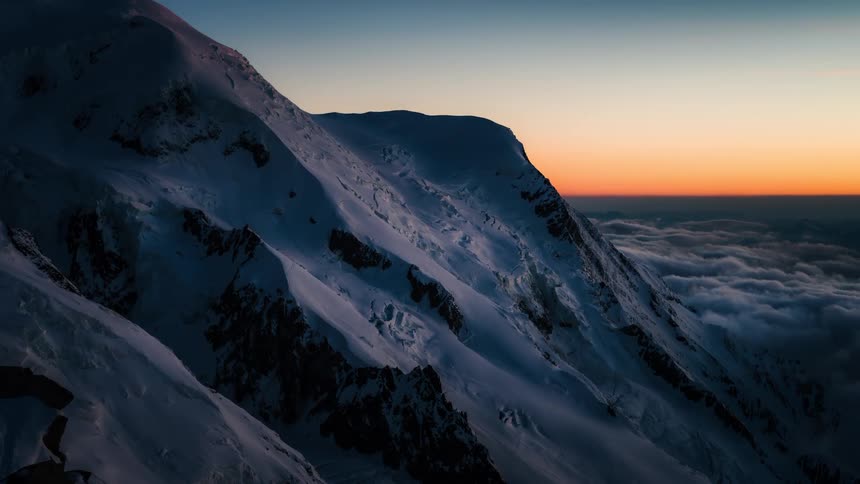 A beautiful timelapse of the sunset casting a warm alpenglow across a majestic snowy mountain range high above a sea of clouds