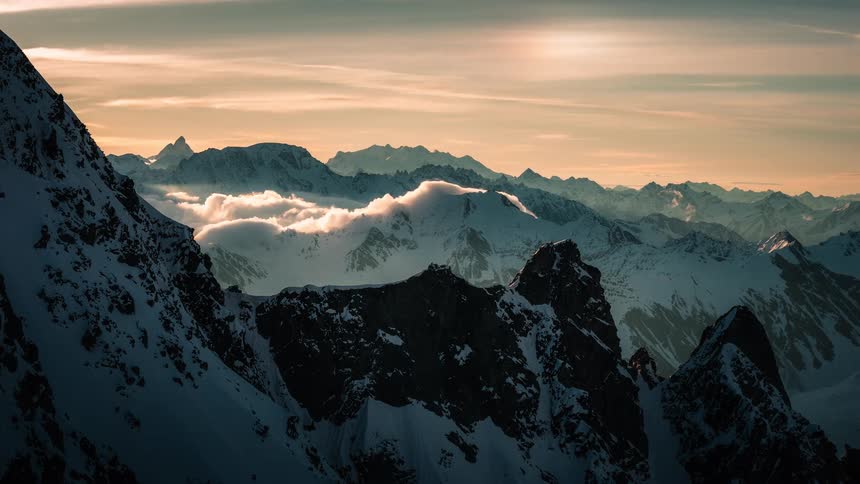 A stunning cinemagraph captures clouds flowing over a majestic, snow-covered mountain range at sunset