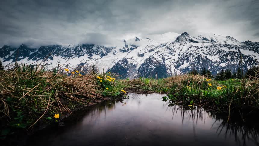 A stunning timelapse captures dramatic clouds rolling over majestic snow-capped mountains, with a serene reflection in a mountain pool surrounded by wildflowers