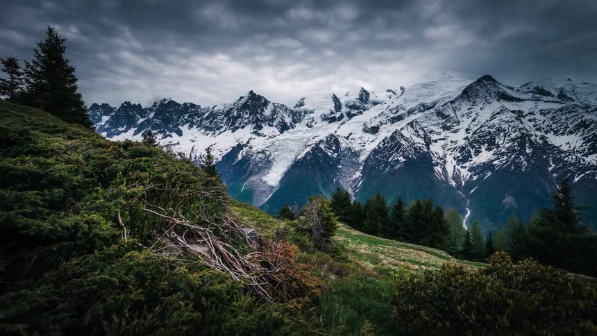 A dramatic timelapse captures moody clouds swirling over the snow-covered peaks of the Mont Blanc massif, viewed from a lush green alpine meadow