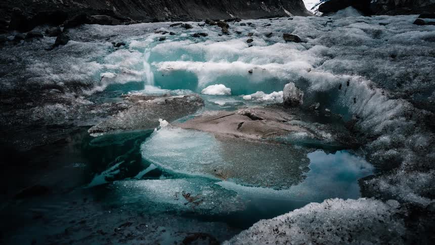 A timelapse of a frozen arctic landscape shows a small waterfall of meltwater flowing over a glacier into a turquoise pool