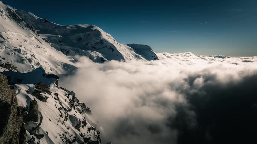 A majestic time-lapse captures a sea of clouds flowing through a snowy mountain range under a clear blue sky