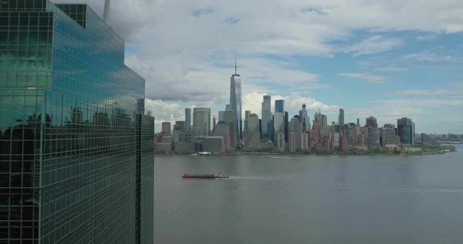 Aerial shot pushes between Jersey City skyscrapers to reveal the iconic Lower Manhattan skyline and a barge on the Hudson River