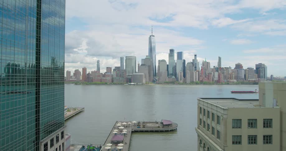 A slow aerial reveal of the New York City skyline and the Hudson River, as seen from between skyscrapers in Jersey City on a partly cloudy day