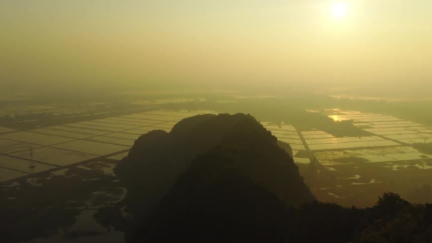 A majestic aerial view shows silhouetted mountains overlooking a vast landscape of flooded rice paddies during a hazy golden sunrise