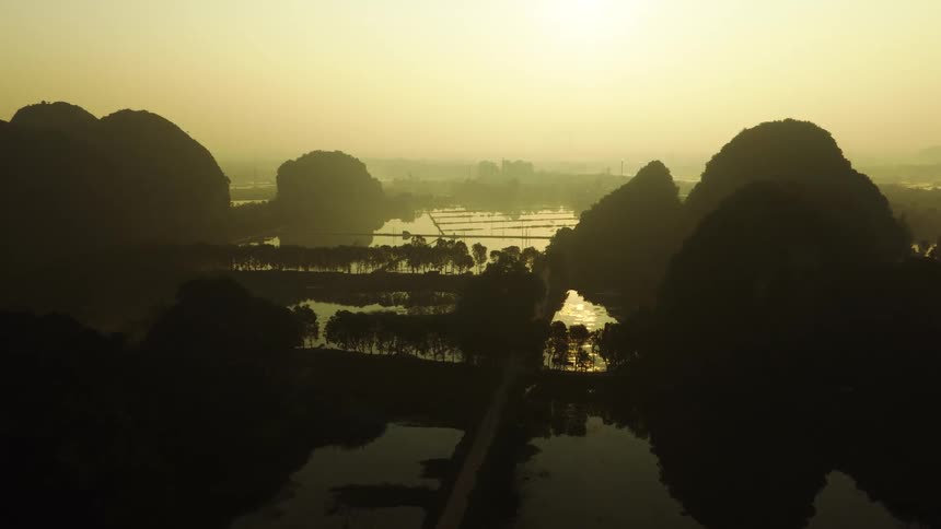 An epic aerial view of silhouetted karst mountains over a tranquil, reflective river valley during a hazy golden hour