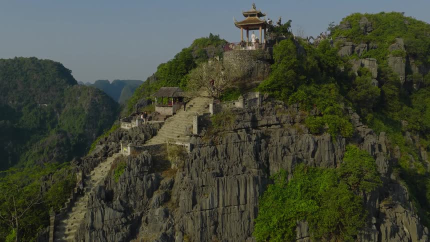 An aerial view rises up a long stone staircase carved into a lush, rocky mountain leading to a scenic pagoda viewpoint in Vietnam