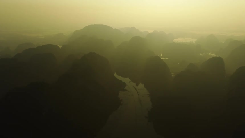 A mystical static aerial view of a river valley and silhouetted mountains shrouded in a thick golden haze at sunrise