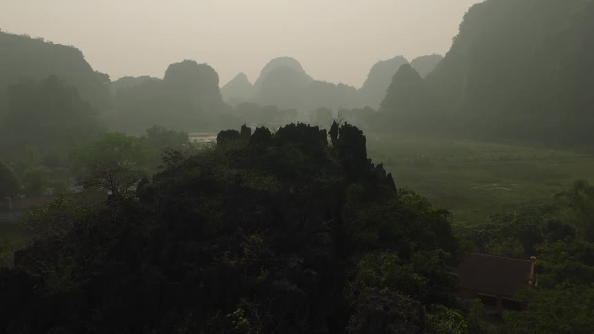An aerial drone slowly flies towards a jagged, jungle-covered mountain peak with a hazy, majestic mountain range in the background