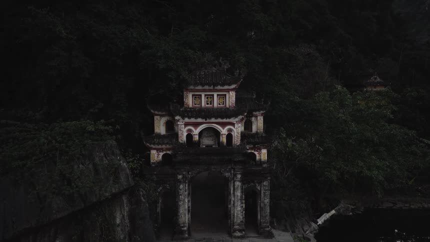 A slow aerial zoom captures an ancient, weathered Vietnamese temple gate nestled in a dark, mysterious forest at dusk