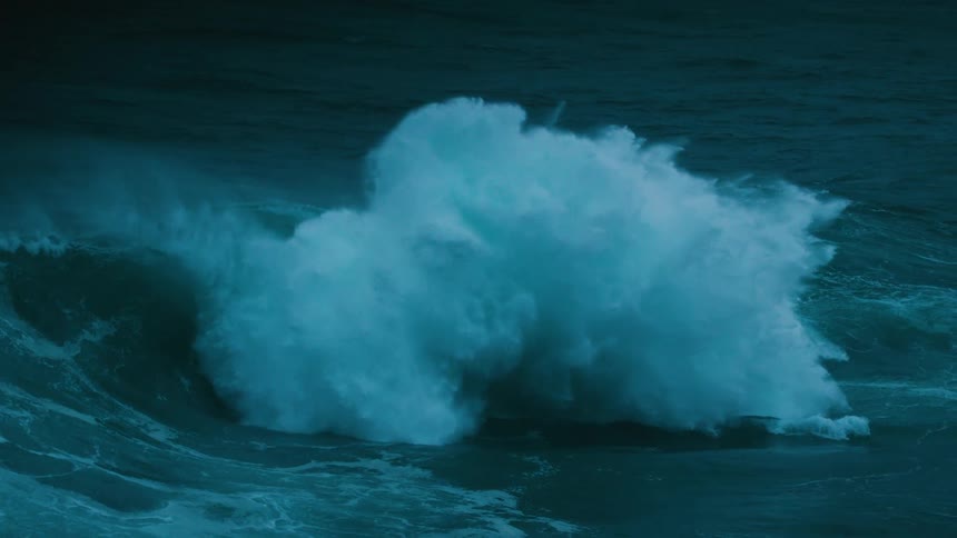 A dramatic slow-motion shot captures the immense power of a large ocean wave breaking in the dark