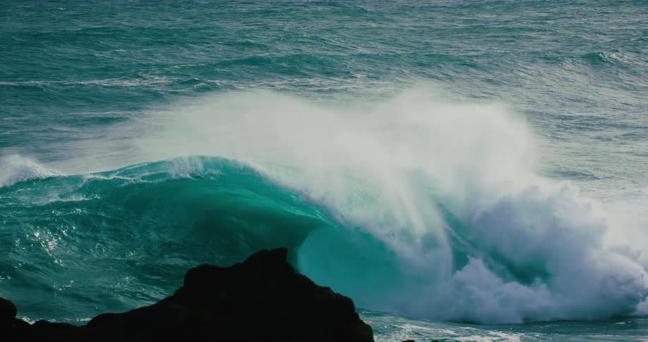 An enormous turquoise ocean wave curls and crashes in dramatic slow motion, viewed from behind silhouetted coastal rocks