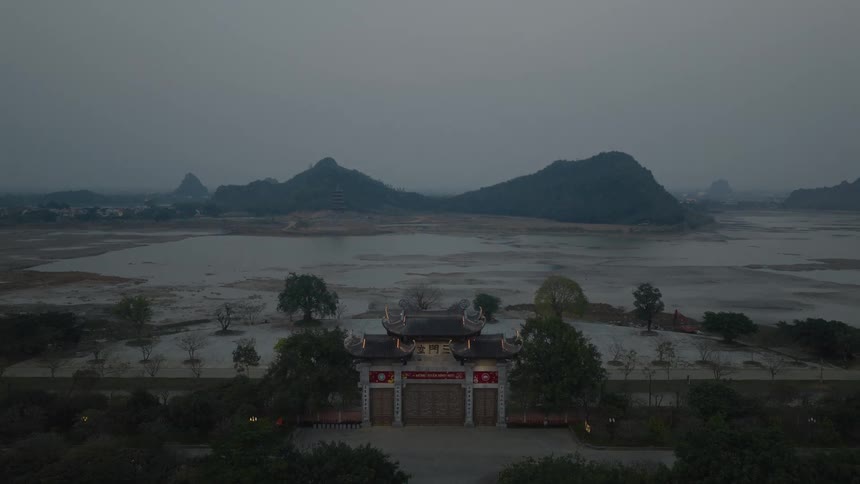 A slow aerial approach to a traditional Vietnamese temple gate at dusk, overlooking a serene lake and mountains