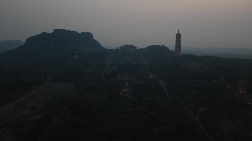 A wide aerial shot of a vast, ancient temple complex and pagoda nestled in a dark, forested mountain range at dusk